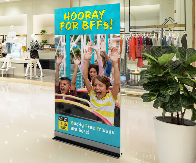 A tall sign that says "hooray for BFFs" and an image of kids on a roller coaster. The sign is sitting in the middle of a mall with a clothing store behind it and a plant to the right of it.