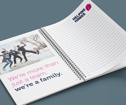 a spiral notebook with an image of three people jumping and "we're more than just a team. We're a family." on the left. Lined pages with the helping hands logo on the right.